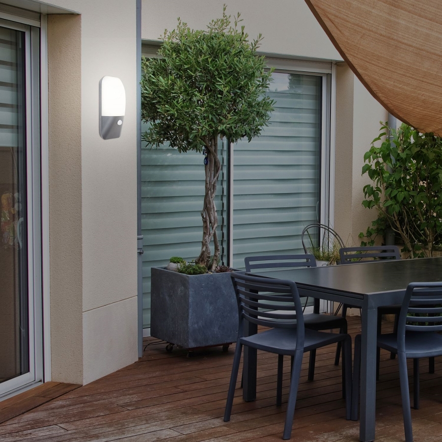 Vonkajšia nástenná lampa svieti. Nachádza sa na exteriéri domu pri jedálenskom stole a rastline v kvetináči. Drevená terasa je okolím.