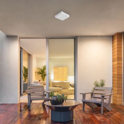 Svietidlo osvetľuje vonkajšiu terasu. Dve drevené stoličky sú umiestnené okolo nízkeho stolíka s kvetináčom. V pozadí sa nachádza presklená stena s výhľadom na interiér domu.