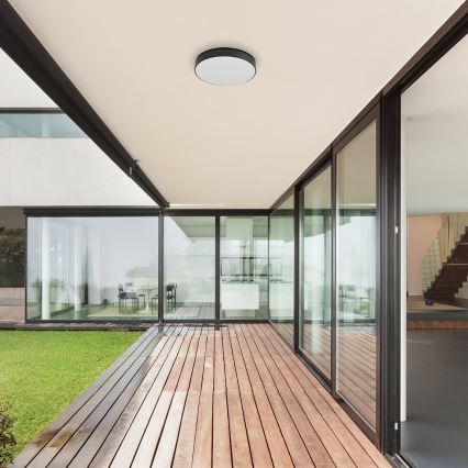 Svetlo svieti na drevenej terase modernej vily. Terasa je pripojená k skleneným stenám s výhľadom na trávnik. Vnútorné priestory vily sú čiastočne viditeľné cez sklenené steny.