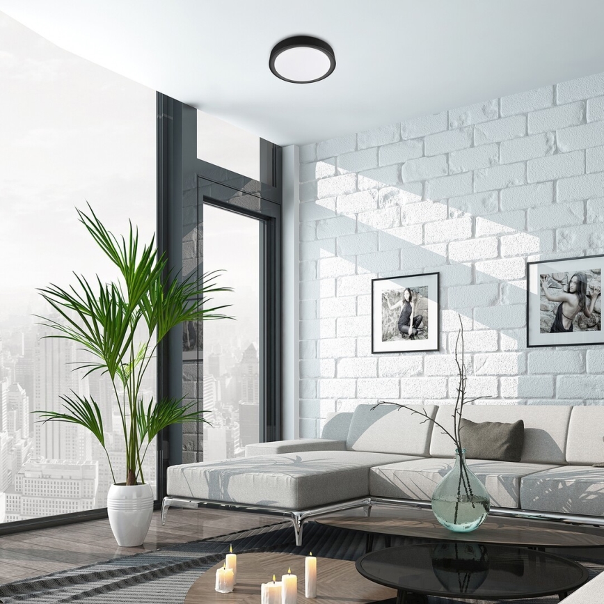 Stropné svietidlo svieti v modernej obývačke. Svetlo je kruhové a tmavosivé. Obývacia izba má bielu tehlovú stenu, sedačku a veľké okno s výhľadom na mesto.