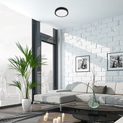 Stropné svietidlo svieti v modernej obývačke. Svetlo je okrúhle, tmavosivé. Obývačka má bielu tehlovú stenu, svetlý nábytok a veľké okno s výhľadom na mesto.
