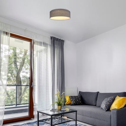 Svietidlo s textilným tienidlom svieti v obývacej izbe. Svietidlo je pripevnené k stropu. Izba obsahuje pohovku, konferenčný stolík a závesy.