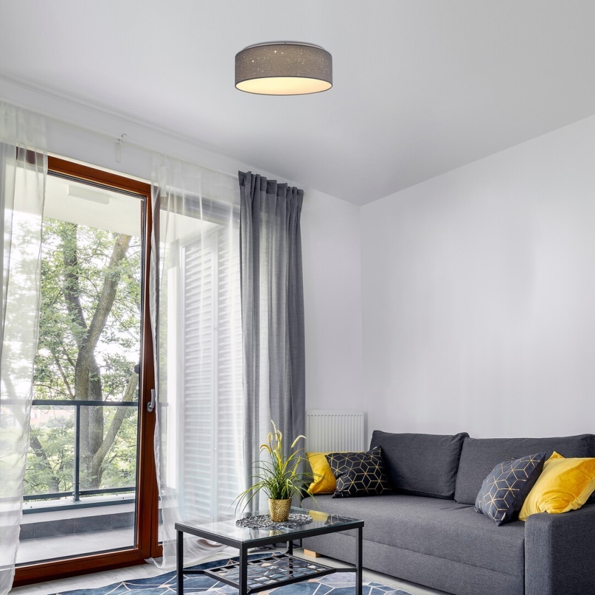Svietidlo s textilným tienidlom svieti v obývacej izbe. Svietidlo je pripevnené k stropu. Izba obsahuje pohovku, konferenčný stolík a závesy.