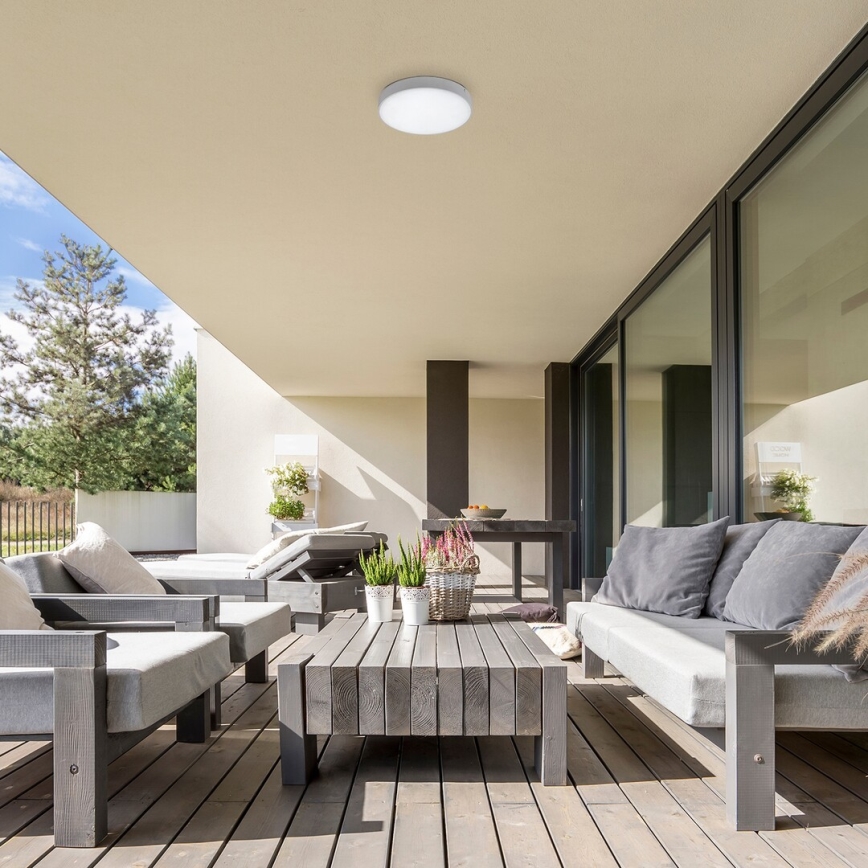 Záhradný stropný svietidlo svieti na terase. Na terase je drevený nábytok: sedačka, kreslá a stolík. Okolo sa nachádzajú rastliny v kvetináčoch.