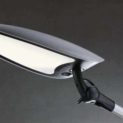 Stolná lampa svieti. Je pripevnená k nastaviteľnému rameni. Pozadie je tmavosivé.