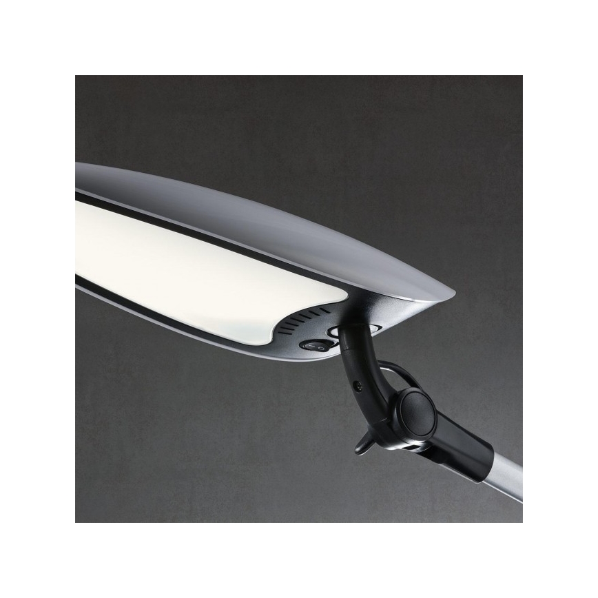 Stolná lampa svieti. Je pripevnená k nastaviteľnému rameni. Pozadie je tmavosivé.