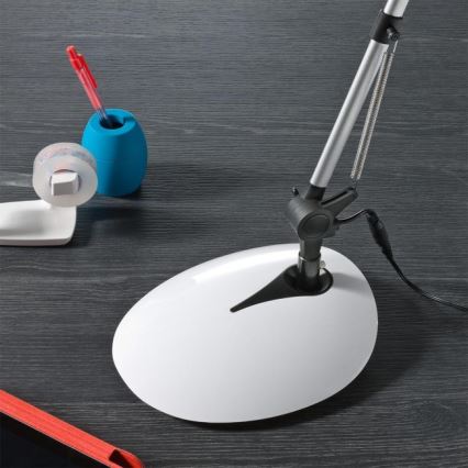 Stolná lampa s bielou základňou a strieborným ramenom stojí na tmavom stole. Vedľa nej je modrá nádobka na pero a náplň lepiacej pásky.