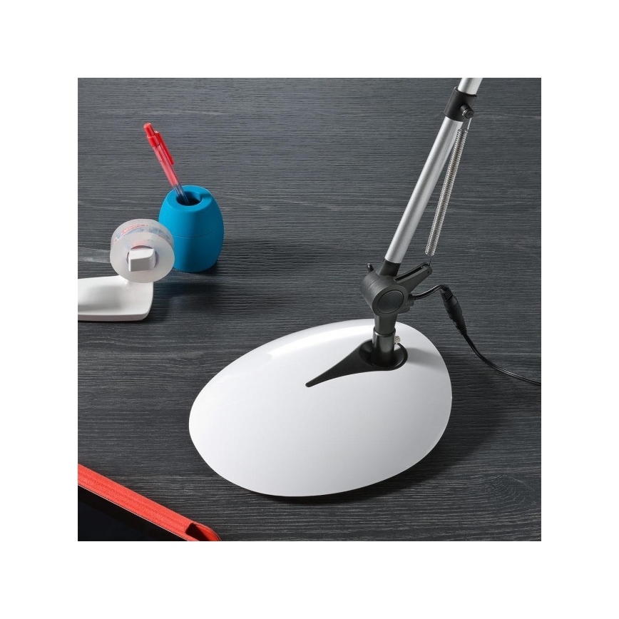 Stolná lampa s bielou základňou a strieborným ramenom stojí na tmavom stole. Vedľa nej je modrá nádobka na pero a náplň lepiacej pásky.