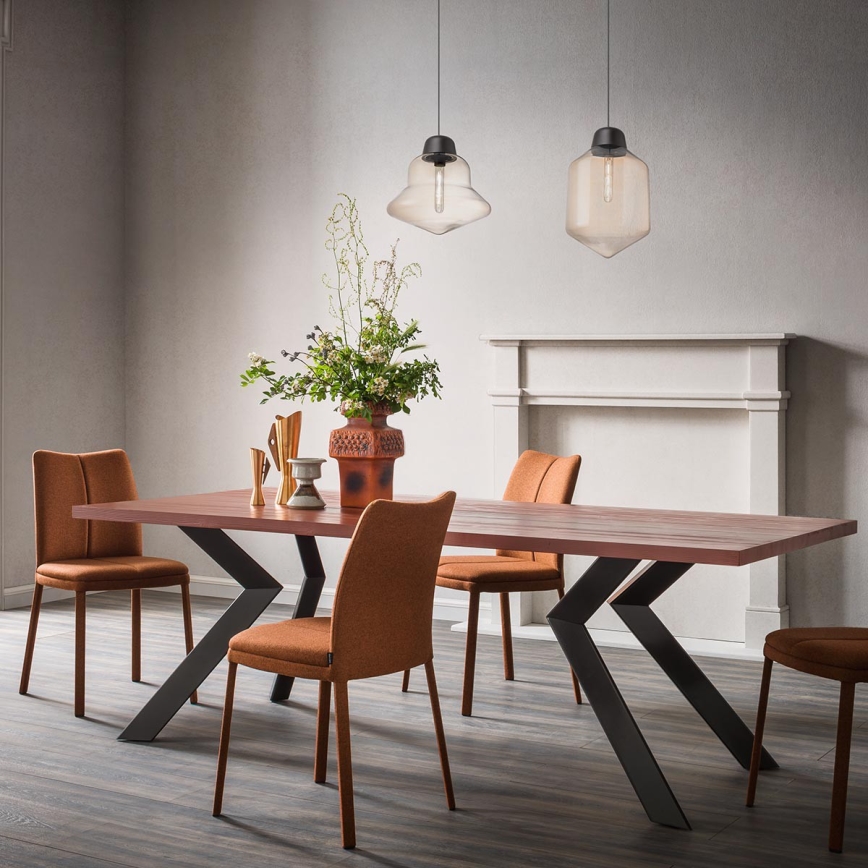 Drevený jedálenský stôl s kovovými nohami v tvare X stojí v jedálni. Okolo stola sú oranžové stoličky. Nad stolom visia dve sklenené závesné lampy. V pozadí je krb.