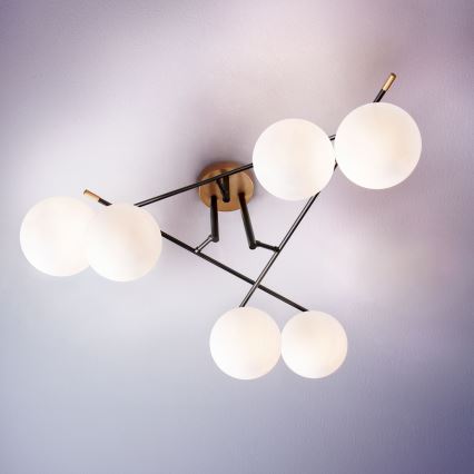 Stropné svietidlo s piatimi guľovými tienidlami z matného skla, pripevnené na čiernom kovovom ráme s zlatými detailmi, je zavesené na svetlofialovom strope.