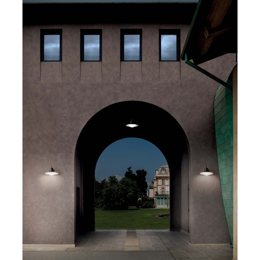 Dve nástenné lampy svietia na oboch stranách klenutej vstupnej brány do tmavofialovej budovy. V pozadí je vidieť záhradu s budovou.