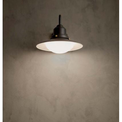 Nástená lampa svieti. Je čierna s bielym, guľovitým tienidlom. Je pripevnená na svetlohnedej stene.