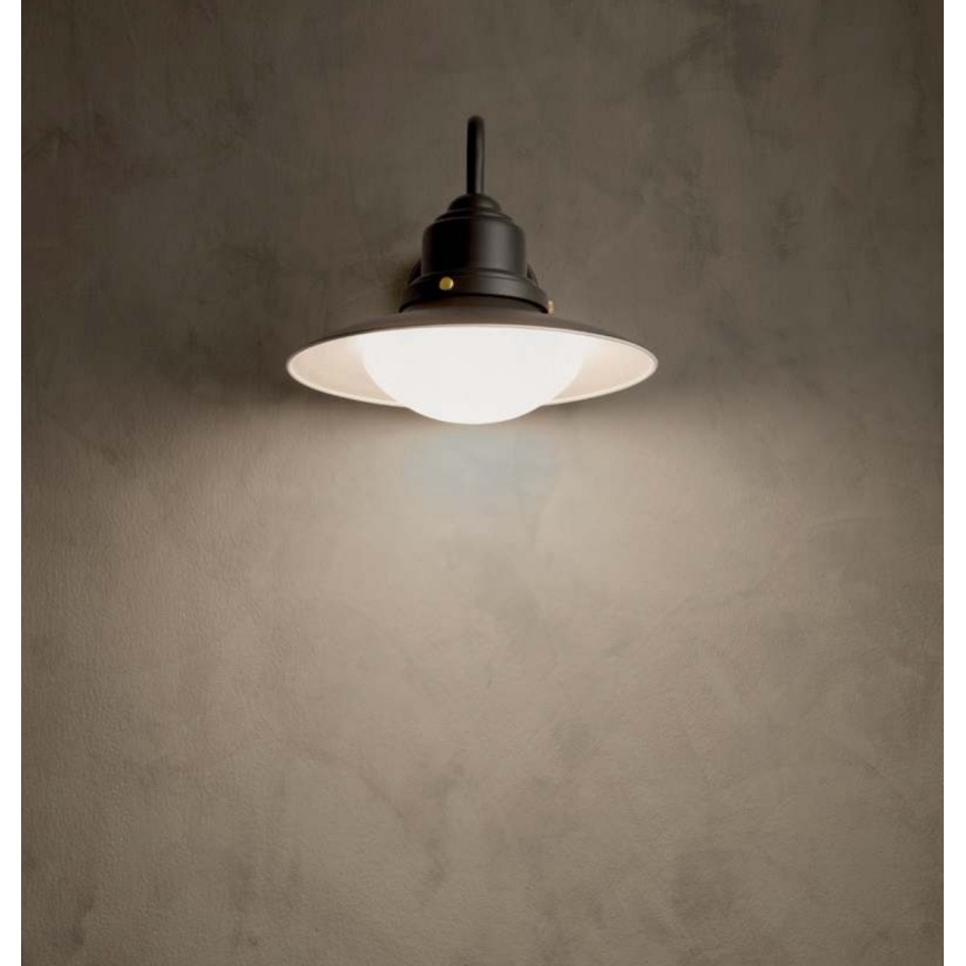 Nástená lampa svieti. Je čierna s bielym, guľovitým tienidlom. Je pripevnená na svetlohnedej stene.