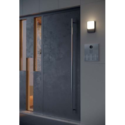 Dvere z tmavého materiálu s kovovou kľučkou sú osvetlené vonkajším svetlom. Vedľa dverí je umiestnený videovrátnik. Celok sa nachádza v modernej exteriérovej architektúre.