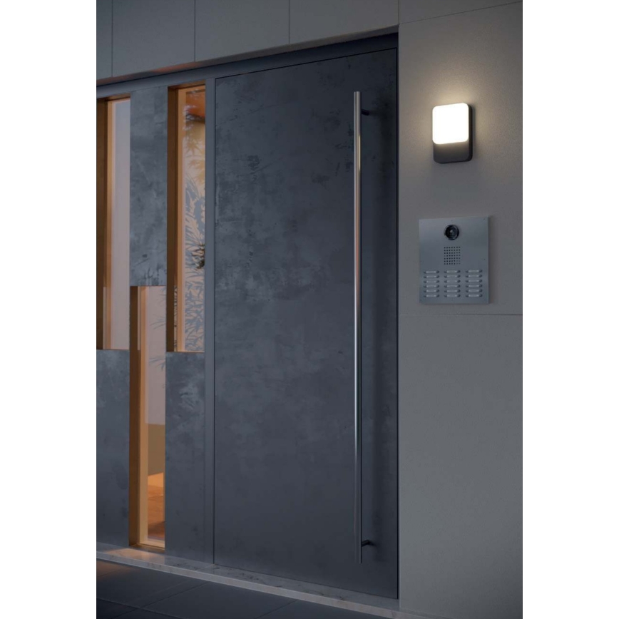 Dvere z tmavého materiálu s kovovou kľučkou sú osvetlené vonkajším svetlom. Vedľa dverí je umiestnený videovrátnik. Celok sa nachádza v modernej exteriérovej architektúre.