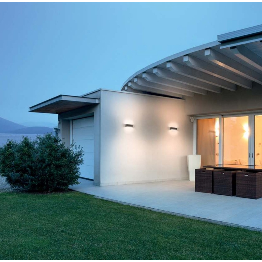 Dve nástenné svietidlá svietia na vonkajšej stene modernej budovy. Budova má plochú strechu s presahom a veľké presklenné dvere. Pred budovou sa nachádza trávnik a terasu s ratanovým nábytkom.