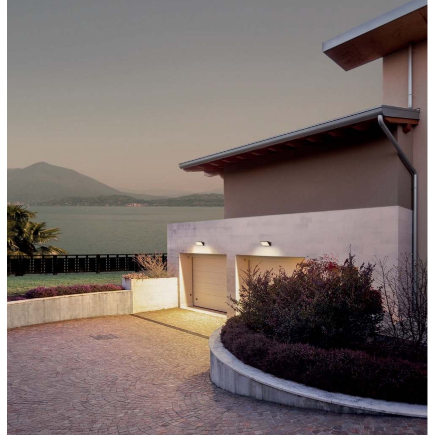 Dve nástenné svietidlá osvetľujú dvojgaráž pri dome. Dom stojí na kopci s výhľadom na jazero a hory.