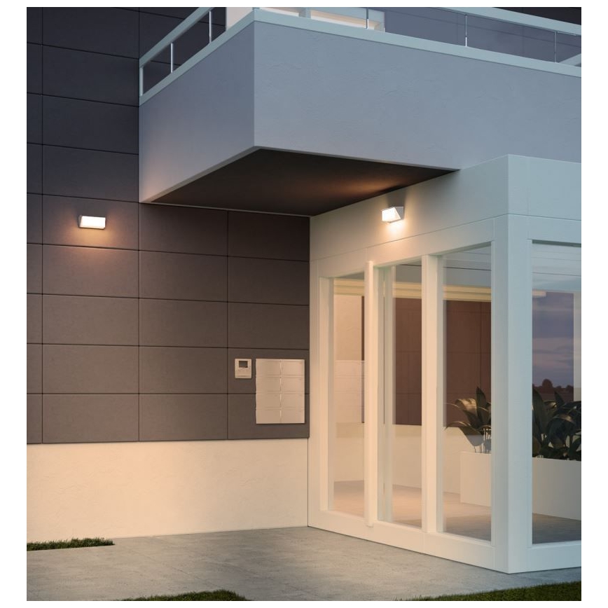 Vonkajšie nástenné svietidlá osvetľujú moderný dom s vonkajším schodiskom a presklenými dverami. Svietidlá sú umiestnené na tmavých obkladačkách. Pri vchode sa nachádza schránka a zvonek.