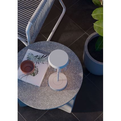 Biela stolová lampa stojí na okrúhlom stolíku z teraca. Na stolíku je aj miska a kniha s obrázkami listov. Stolík sa nachádza na terase s dlažbou.