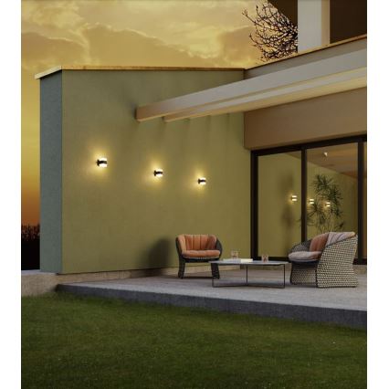 Tri vonkajšie nástenné svietidlá svietia na zelenej stene domu. Pred stenou je terasa s dvoma kreslami a stolíkom. Trávnik sa nachádza pred terasou.