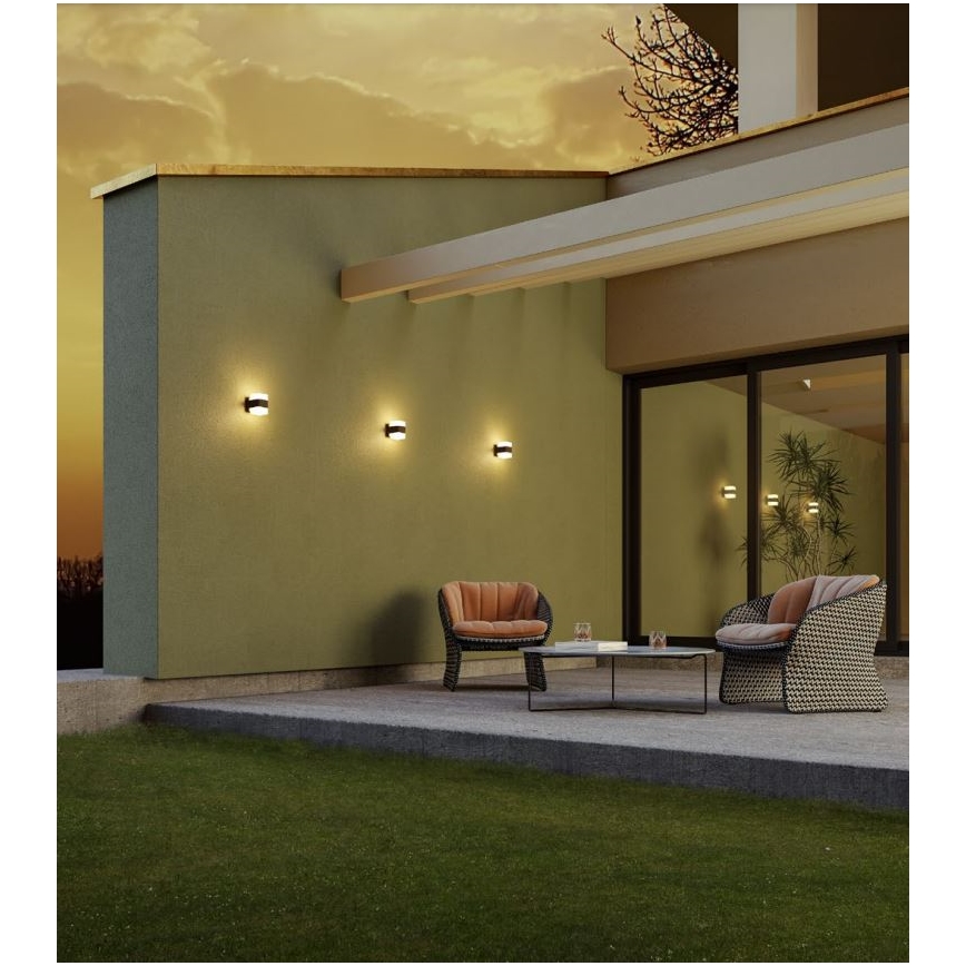 Tri vonkajšie nástenné svietidlá svietia na zelenej stene domu. Pred stenou je terasa s dvoma kreslami a stolíkom. Trávnik sa nachádza pred terasou.