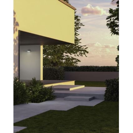 Vonkajšie nástenné svietidlo je namontované na svetlej stene modernej budovy. Svietidlo osvetľuje schody a chodník vedúci k domu. Okolie tvorí trávnik, nízky ker a stromy za súmraku.