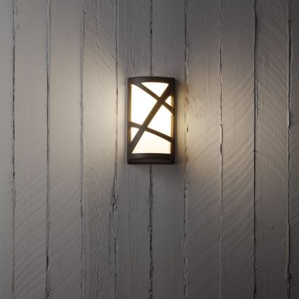 Svietidlo svieti na stene. Obdĺžnikové svietidlo z tmavého kovu s mliečnym sklom a vnútornými ozdobnými líniami je pripevnené k svetlej drevenej stene.