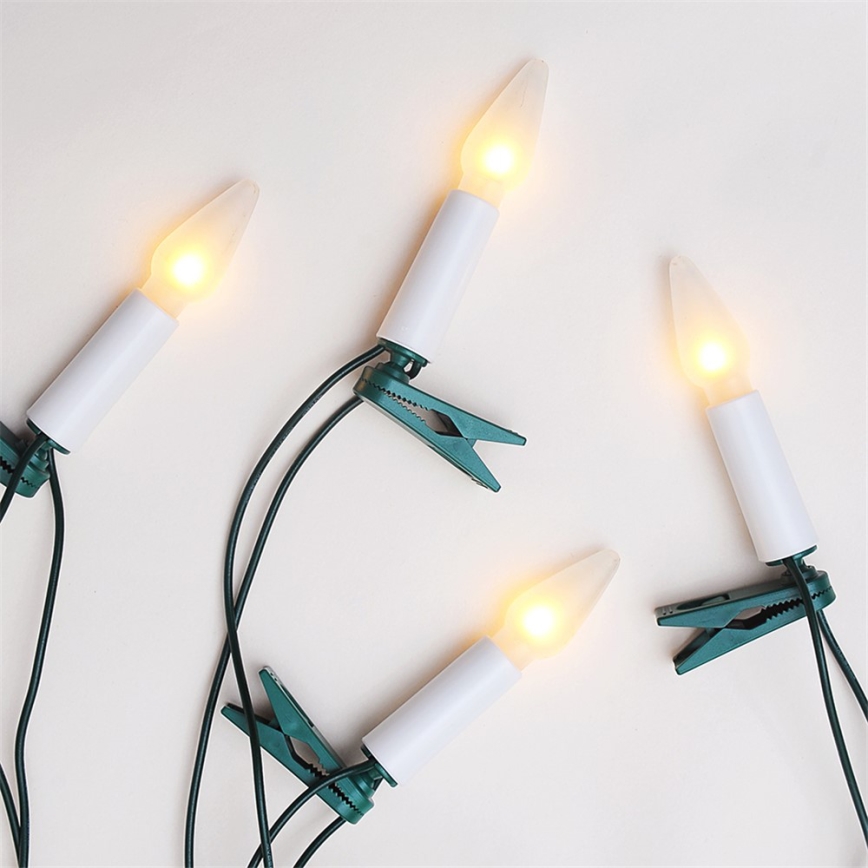 Štyri LED sviečky pripevnené zelenými sponkami na kábel. Sviečky sú biele s teplým žltým svetlom. Svetlá sú umiestnené na svetlom pozadí.