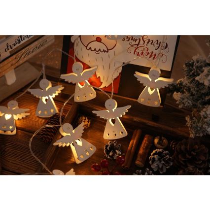 Svetlá vo forme drevených anjelikov s vyrezávanými srdiečkami svietia. Sú umiestnené na drevenom podnose s vianočnými dekoráciami ako šišky a škorica. Na pozadí je vianočná pohľadnica s nápisom Merry Christmas.