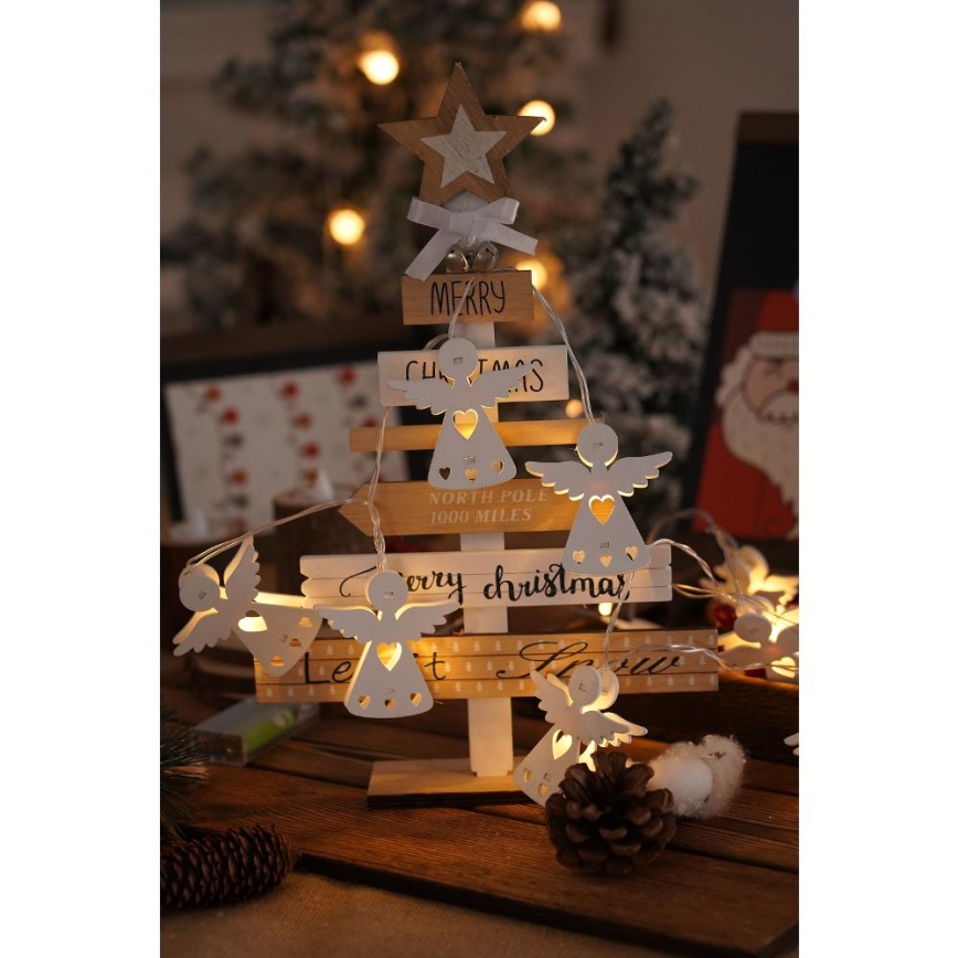Drevený vianočný stromček s LED svetielkami v tvare anjelikov svieti. Na stromčeku sú drevené tabuľky s nápismi: „MERRY CHRISTMAS“, „NORTH POLE 1000 MILES“, „Merry christmas“. Stromček stojí na drevenom povrchu s vianočnými ozdobami.