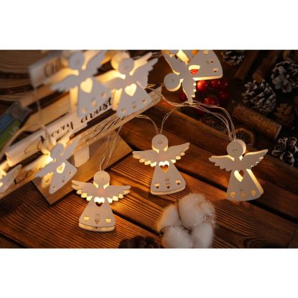 Svetlá vo forme drevených anjelikov s vyrezávaným srdiečkom svietia na drevenom stole. Anjeliky sú zavesené na svetelnom reťazi. Na pozadí je drevený stojan s nápisom „Merry Christ...“.