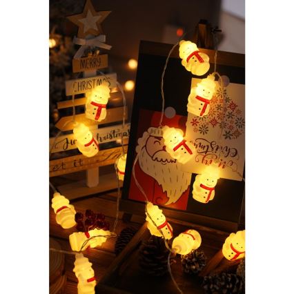 Svetielka v tvare snehuliakov svietia. Sú umiestnené na šnúre a rozložené na drevenom povrchu s vianočnými dekoráciami, vrátane dreveného stromčeka s nápisom Merry Christmas a obrázka Santa Clausa.