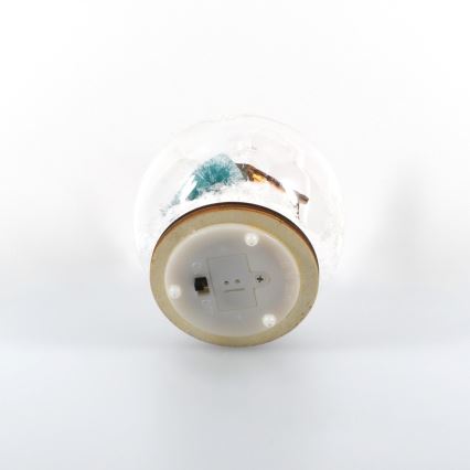 Snehová guľa s vianočnou scénkou je položená na bielom podklade. Viditeľný je spodný drevený kryt s vypínačom.