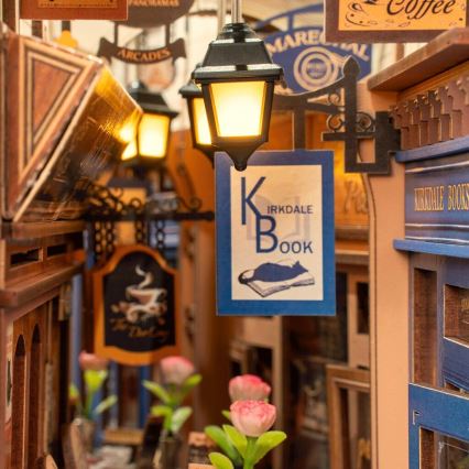 Miniatúrna ulička s obchodmi; osvetlená lampami; na značke kníhkupectva „Kirkdale Book“ je mačka ležiaca na knihe.