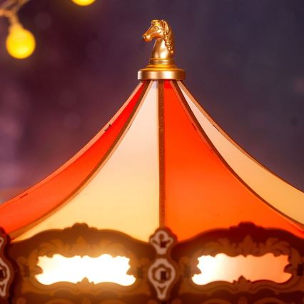 Miniatúrny kolotoč s červeno-bielym stanom a zlatým koňom na vrchu svieti v rozostrenom pozadí s rozmazanými svetlami.