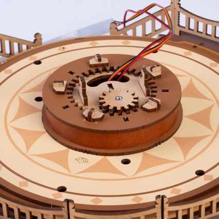 Drevená ozubená sústava sa otáča v strede kruhovej plošiny s ozdobným okrajom. Súčasťou je aj vonkajší drevený rám a káble.