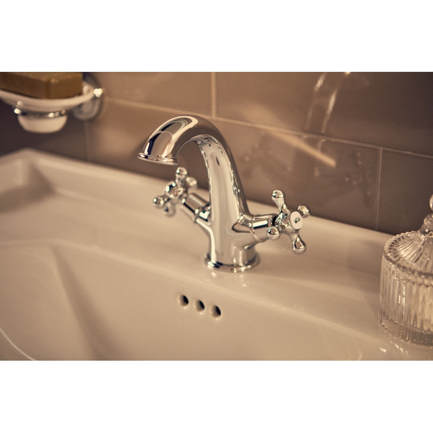 Dvojica chrómovaných batériových pák sa nachádza na keramickom umývadle v kúpeľni s hnedými obkladmi. V pozadí je viditeľný držiak na mydlo.