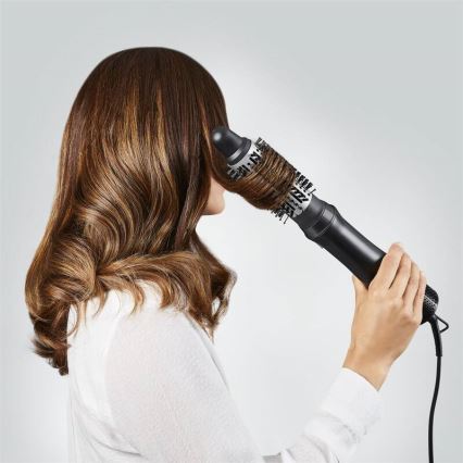 Žena s dlhými hnedými vlasmi používa kulmu s okrúhlou kefou na úpravu vlasov. Kefou si tvaruje vlasy pred svetlosivým pozadím.