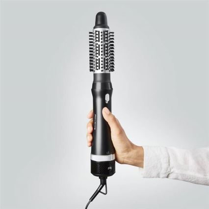 Rotačná kefová žehlička na vlasy s čiernym telom a striebornými prvkami drží ruka. Kefa má valcový tvar s štetinami. Prístroj je pripojený káblom k elektrickej sieti.