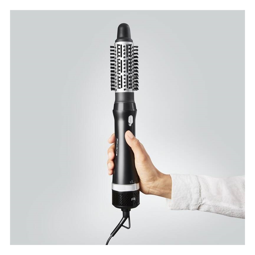 Rotačná kefová žehlička na vlasy s čiernym telom a striebornými prvkami drží ruka. Kefa má valcový tvar s štetinami. Prístroj je pripojený káblom k elektrickej sieti.