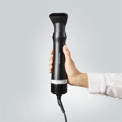 Ruka drží čierny fén Rowenta s nástavcom. Fén má strieborný prvok a sieťku na spodnej časti. Na pozadí je svetlosivá stena.