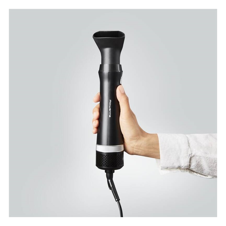 Ruka drží čierny fén Rowenta s nástavcom. Fén má strieborný prvok a sieťku na spodnej časti. Na pozadí je svetlosivá stena.