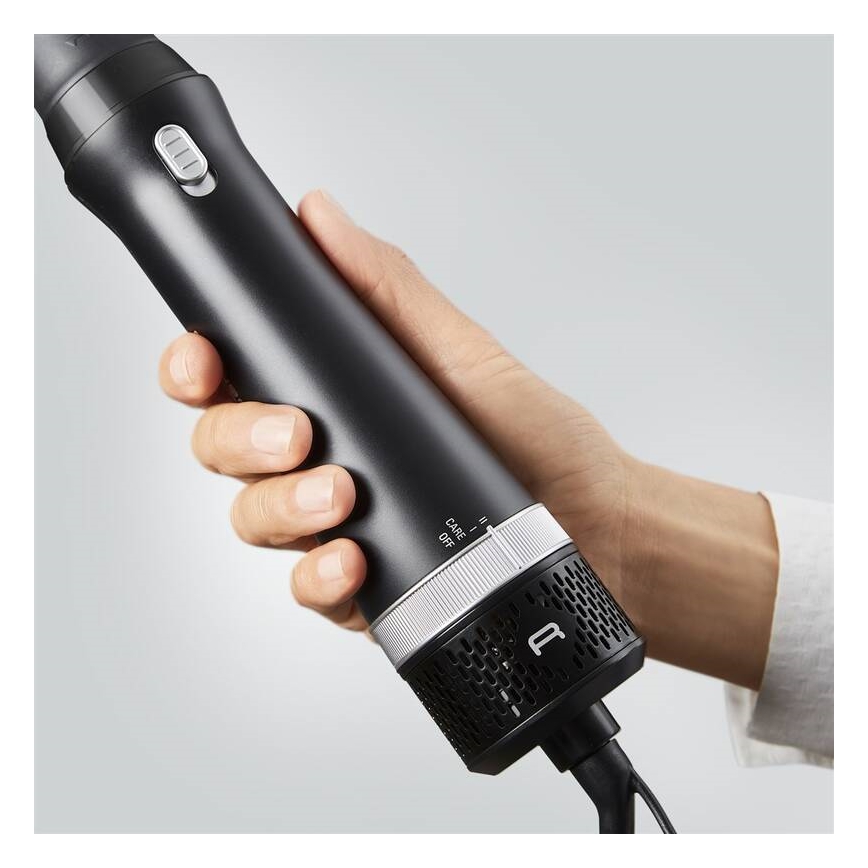 Ruka drží čierny fén s kovovými prvkami. Na féne je otočný volič s označením „CASE - II - OFF“. Fén má štrbiny na spodnom konci.