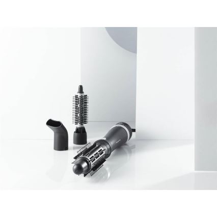 Fén s nástavcami leží na bielom povrchu. Súčasťou je hlavica s okrúhlou kefou a ďalšie dve menšie nástavce, jeden s dlhými a jeden s krátkymi štetinami. V pozadí sú biele steny.