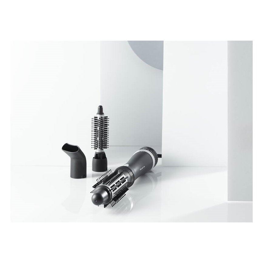 Fén s nástavcami leží na bielom povrchu. Súčasťou je hlavica s okrúhlou kefou a ďalšie dve menšie nástavce, jeden s dlhými a jeden s krátkymi štetinami. V pozadí sú biele steny.