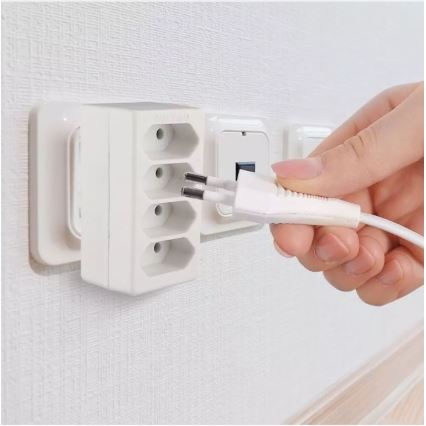 Biela zásuvková predlžovačka s troma otvormi sa pripája do elektrickej zásuvky. Ruka zasúva do nej bielu elektrickú zástrčku. Predlžovačka je namontovaná na stene.