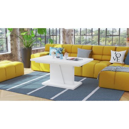 Biely konferenčný stolík s rozkladacou doskou stojí v obývacej izbe pred žltou rozkladacou sedačkou. Na stolíku sú dekorácie. Stolík je umiestnený na tmavomodrom koberci s bielymi pruhmi.