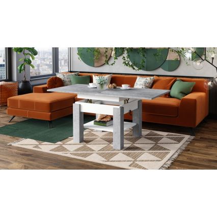 Rozťahovací konferenčný stolík so šedou doskou a bielymi nohami stojí pred oranžovou sedačkou v obývačke. Stolík má jednu poličku a je umiestnený na koberci s geometrickým vzorom.