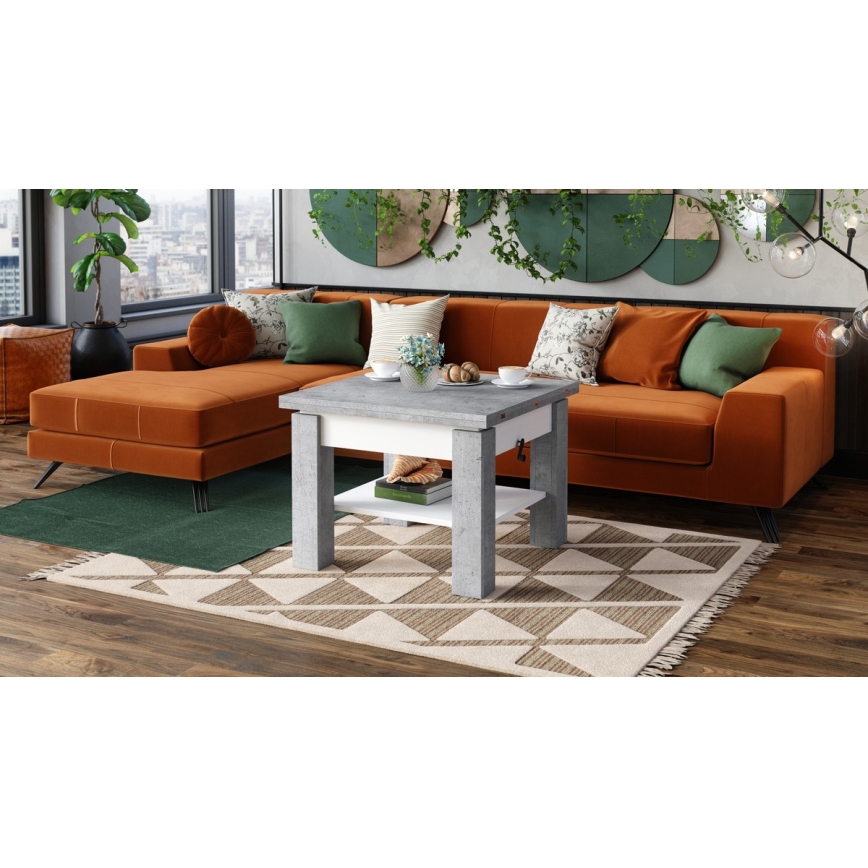 Odkládací stolík so šuflíkom a poličkou stojí pred rohovou sedačkou v obývacej izbe. Stolík je sivý s bielou vnútornou časťou. Sedačka je oranžovej farby, izba má drevenú podlahu a koberec s geometrickým vzorom.