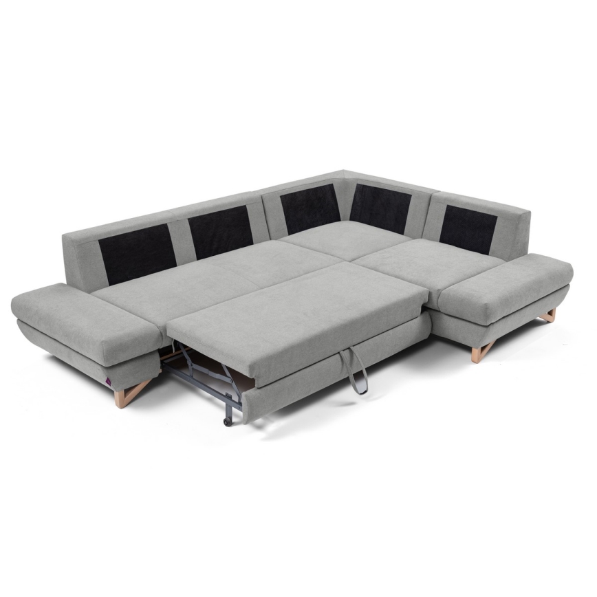 Rohová sedačka s rozkladacím mechanizmom sa rozkladá. Sedačka je sivá s čiernymi detailmi, má drevené nožičky a je umiestnená na bielom pozadí.